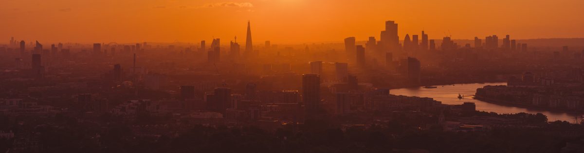 Panoramic aerial view of the London skyline at sunset, with The Shard, City of London and River Thames silhouetted through golden atmospheric light.